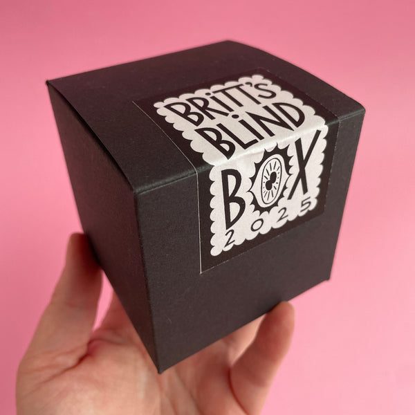 Black cube box 3x3x3inches, held in a hand on a pink background with a black and white sticker featuring ahnd lettering that says "BRITT'S BLIND BOX 2025" but the O in box is replaced by a very stylized eyeball with 2 pupils.