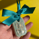 A hand holding a small short rectangle ceramic tile ornament decorated with an aqua crackled glaze and golden hand drawn ribbon