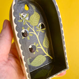 An arched ceramic wall shelf featuring an illustration of a stylized Atropa Belladonna plant. Held in a hand for scale.