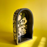 An arched ceramic wall shelf featuring an illustration of stylized cartoon skulls and Dandelion plants. a Front view
