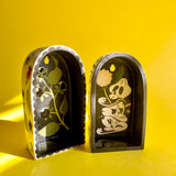 Two arched ceramic wall shelves, one with an illustration of Atropa Belladonna berries and flowers, and the other with a pile of cartoon skulls and dandelion bits coming out of them.