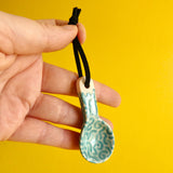 A hand holding a small ceramic spoon decorated with an aqua coloured glaze and turquoise coloured wiggly worm shapes.